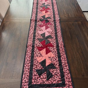 May include: A quilted table runner with a pinwheel design. The runner features a repeating pattern of pinwheels in shades of pink, red, and black, bordered by a black frame. The runner is placed on a dark wooden table.