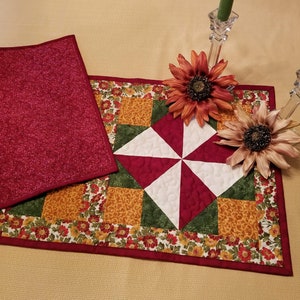 Pu&ograve; includere: Una tovaglia trapuntata con un motivo a ruota al centro, con quadrati rossi, bianchi, verdi e oro. La tovaglia &egrave; bordata con un motivo floreale e un bordo rosso pieno. Un pezzo di tessuto rosso abbinato &egrave; accanto alla tovaglia.