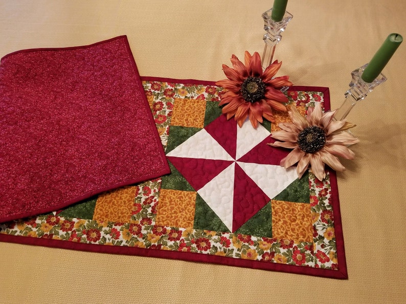 Pu&ograve; includere: Una tovaglia trapuntata con un motivo a ruota al centro, con quadrati rossi, bianchi, verdi e oro. La tovaglia &egrave; bordata con un motivo floreale e un bordo rosso pieno. Un pezzo di tessuto rosso abbinato &egrave; accanto alla tovaglia.