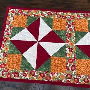 Pu&ograve; includere: Un runner da tavolo trapuntato rettangolare con bordo rosso. Il centro presenta un motivo a mulinello in bianco e rosso, circondato da quadrati verdi e arancioni. I bordi sono adornati da un motivo floreale rosso, giallo e arancione.