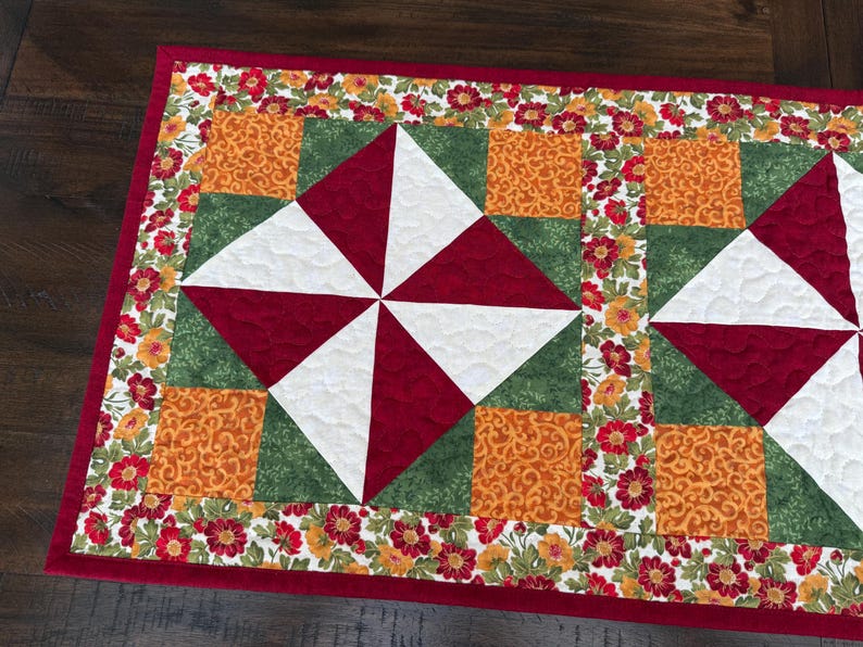 Pu&ograve; includere: Un runner da tavolo trapuntato rettangolare con bordo rosso. Il centro presenta un motivo a mulinello in bianco e rosso, circondato da quadrati verdi e arancioni. I bordi sono adornati da un motivo floreale rosso, giallo e arancione.