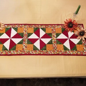 May include: A rectangular quilted table runner with a repeating pattern of red, white, and green geometric shapes. The runner is bordered with a floral print and is decorated with artificial sunflowers and candle holders.