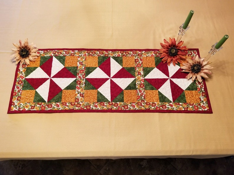 Pu&ograve; includere: Una tovaglia rettangolare trapuntata con un motivo ripetuto di forme geometriche rosse, bianche e verdi. La tovaglia &egrave; bordata con una stampa floreale ed &egrave; decorata con girasoli artificiali e candelabri.