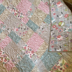 May include: A patchwork quilt with a floral and owl design. The quilt is made of various shades of pink, brown, and blue fabric. The quilt is quilted with a swirl pattern.
