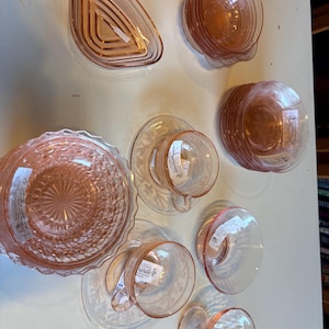 May include: A collection of vintage pink depression glass tableware including a triangular dish, a scalloped bowl, a stack of small bowls, two teacups with matching saucers featuring floral patterns, and a stemmed glass, all with white price tags.
