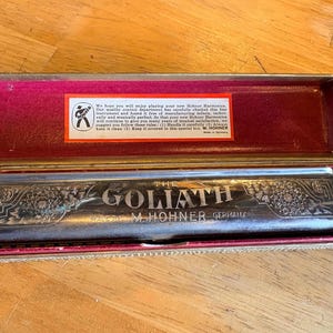 May include: A silver-toned metal harmonica labeled The Goliath and M. Hohner Germany, featuring engraved floral patterns. It is housed in a rectangular case with a red interior lining and a white paper label containing printed text instructions.