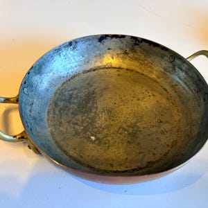May include: A round, shallow metal pan with a weathered, dark interior surface and two curved brass handles attached to the sides. The exterior of the pan shows a warm copper tone, while the interior displays signs of wear and discoloration.