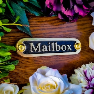 May include: A rectangular mailbox plaque with the word "Mailbox" in white lettering on a black background. The plaque has a gold-colored border and gold accents on each end. The plaque is surrounded by flowers and greenery.