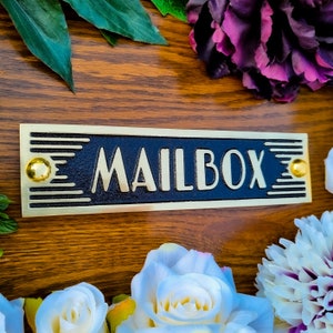 May include: A gold mailbox sign with black lettering that reads "MAILBOX". The sign has a decorative design with gold accents and is mounted on a wooden surface.