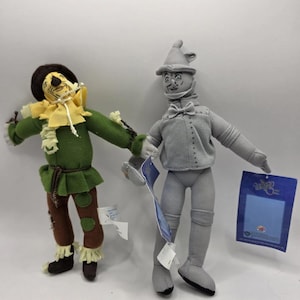 Op de afbeelding: Twee pluche figuren van personages uit The Wizard of Oz. De Vogelverschrikker draagt een groene outfit met een strohoed. De Blikken Man is grijs met een puntmuts en een vlinderdas. Beide figuren hebben labels.