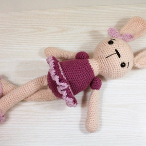 May include: A crocheted bunny doll wearing a pink and purple dress. The doll has black eyes and a pink bow on its head.