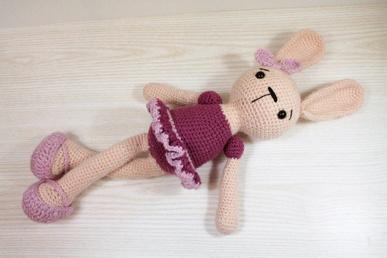 May include: A crocheted bunny doll wearing a pink and purple dress. The doll has black eyes and a pink bow on its head.