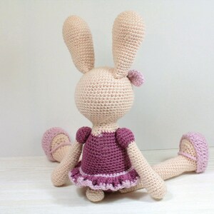 May include: A crocheted bunny wearing a pink and purple dress. The bunny is sitting on a white surface.