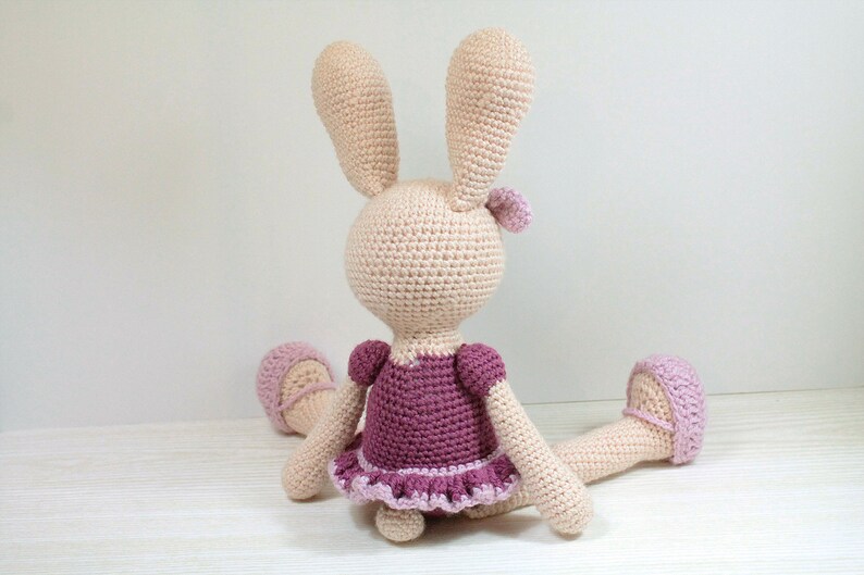 May include: A crocheted bunny wearing a pink and purple dress. The bunny is sitting on a white surface.