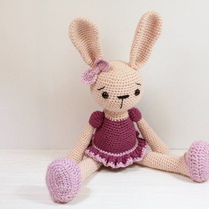 May include: A crocheted pink bunny wearing a purple dress with a pink flower on its head. The bunny is sitting on a white surface.