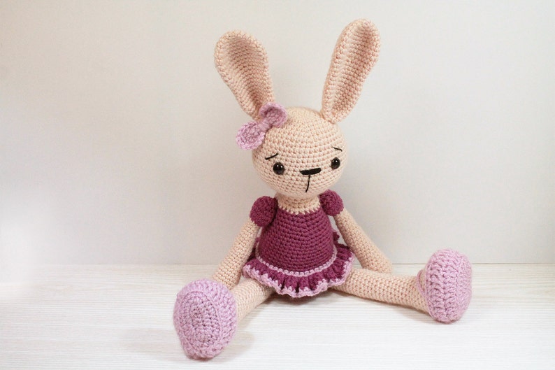 May include: A crocheted pink bunny wearing a purple dress with a pink flower on its head. The bunny is sitting on a white surface.