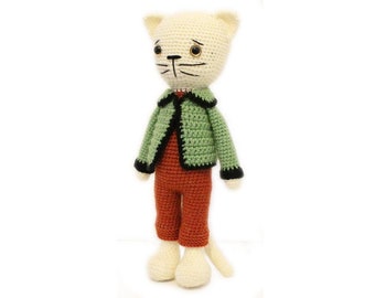 May include: A crocheted cat doll wearing a green jacket with black trim and orange pants. The cat has white fur and black eyes.