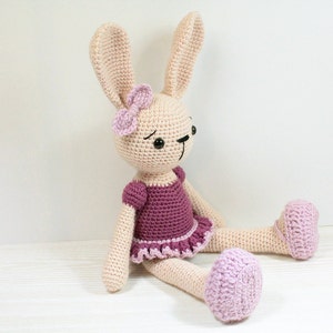 May include: A crocheted bunny wearing a purple dress with a pink bow. The bunny has black eyes and a pink nose. The bunny is sitting on a white surface.