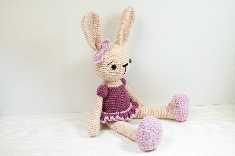 May include: A crocheted bunny wearing a purple dress with a pink bow. The bunny has black eyes and a pink nose. The bunny is sitting on a white surface.