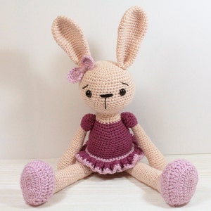 May include: A crocheted pink bunny wearing a purple dress with a pink flower on its head. The bunny is sitting on a white surface.