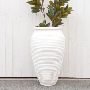 May include: White ceramic planter with a ribbed texture, featuring a green leafy plant inside. The planter is tall and narrow, with a rounded base and a slightly flared top.