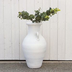 May include: A white ceramic vase with a handle, filled with green leafy branches against a white wood background.