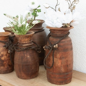 May include: A collection of wooden vases with a rustic aesthetic. The vases are various sizes and feature a warm brown color, with rope detailing around the neck. Some vases hold white flowers and greenery, adding a natural touch.