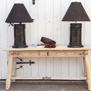 May include: Two black lamps with black lampshades sit on a light wood console table with carved details. The table has a black metal hinge on the side. A small red box and a book are on the table between the lamps.