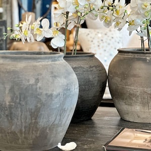 May include: Three gray, textured ceramic vases of varying sizes. The vases are round with wide openings and sit on a dark wooden surface. White orchid flowers with yellow centers are in two of the vases. A decorative home decor item.
