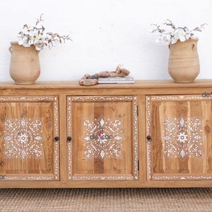 May include: A wooden sideboard with three doors, featuring white floral designs. Two vases with white flowers sit on top, along with decorative objects. The sideboard is made of light-colored wood.