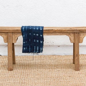May include: A rustic wooden bench with a dark blue patterned textile draped across it. The bench has a natural wood finish and a simple, yet elegant design. The textile adds a pop of color and texture to the scene. The bench is placed on a woven rug.