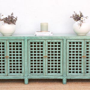 May include: A distressed turquoise cabinet with four doors and a grid pattern. Two white vases with dried flowers sit on either end, with a candle and books in the center. The cabinet is on a woven rug.