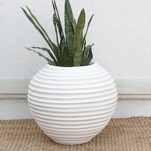 May include: A white, spherical planter with a textured, ridged design. The planter holds a green snake plant. The planter is sitting on a woven, tan rug. Home decor.