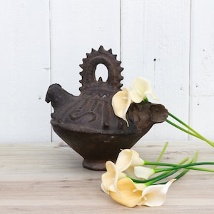 May include: A dark brown ceramic pitcher with a unique design and a handle shaped like a bird. The pitcher is decorated with intricate carvings and is sitting on a wooden surface. A bouquet of white calla lilies with green stems is lying next to the pitcher.