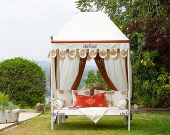 May include: A white outdoor canopy daybed with a brown trim and decorative scalloped valance. The daybed features cream-colored curtains, decorative pillows, and a red rectangular pillow. The canopy has the word "defcor" printed on it.