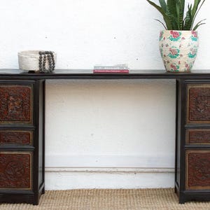 May include: A dark wood console table with ornate, carved panels in a reddish-brown hue. The table has a long, rectangular top with a decorative basket, books, and a floral-patterned vase with a plant. The table is set against a white wall.