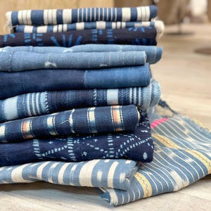 May include: A stack of folded textiles featuring various shades of blue and white. The fabrics display diverse patterns including stripes, geometric shapes, and ikat-style designs, with some pieces showing frayed edges and visible stitching details.