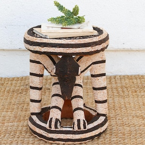 May include: A cylindrical stool covered in intricate beadwork featuring alternating horizontal stripes of cream and dark brown. The base and top are circular, connected by three structural legs that meet in the center, topped with two books and a green plant.