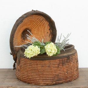 May include: A round, woven basket with a lid, featuring a natural brown color. The lid is partially open, revealing a circular wood grain pattern. The basket is adorned with light green and silver-colored flowers, creating a rustic, decorative piece.