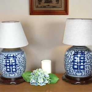 May include: Two blue and white porcelain table lamps with white linen lampshades. The lamps feature a traditional Chinese design with the characters "Double Happiness" in blue.