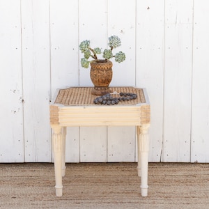 May include: A small, cream-colored side table with a woven top and carved legs. A carved wooden vase with green succulents and a string of dark beads rest on the table. The table is set against a white wooden wall.
