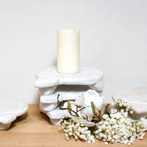 May include: Three white marble candle holders, one stacked on top of the other, with a white candle on the top holder. A bouquet of white flowers is in front of the candle holders.