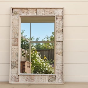 May include: Rectangular mirror with a carved, light-colored wooden frame. The frame has a repeating pattern of carved squares. The mirror reflects a view of a garden with white flowers and a blue sky. The mirror is on a wooden surface.