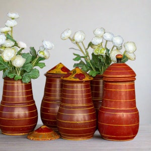 May include: Five wooden containers with lids, painted in red and yellow stripes with floral designs. The containers are arranged in a row on a white surface. Two white flower arrangements are in the background.