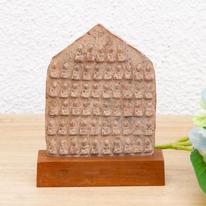 May include: A small, rectangular, brown wooden plaque with a raised design of many small figures. The plaque is on a wooden base and has a light brown color.