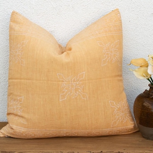 May include: A square mustard yellow throw pillow featuring white embroidered geometric patterns and a border design. Next to the pillow is a dark brown ceramic vase containing a bouquet of light yellow and white calla lily flowers.