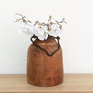 May include: A rustic, wooden vase with a dark brown finish, holding white flowers. The vase has a handle made of a dark, woven material. The flowers are white with yellow centers. The vase is sitting on a wooden surface.