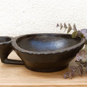 May include: A dark brown wooden bowl with a unique design, featuring a large bowl and a smaller attached cup. The bowl has a handle and is placed on a wooden surface. Decorative greenery is placed to the right of the bowl.