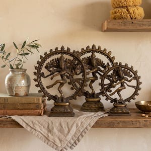 May include: Three bronze-colored metal statues of the deity Shiva Nataraja, each depicted in a dancing pose within a circular flame-like ring, mounted on rectangular bases. The figures are arranged in a row, varying in size from largest to smallest.