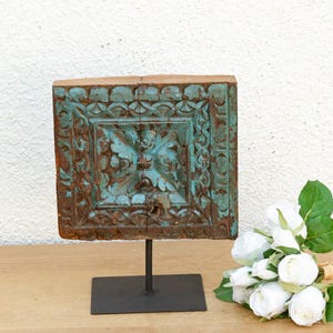 May include: A square, antique wooden panel with intricate carvings, painted in turquoise and brown, mounted on a black metal stand. A bouquet of white roses rests nearby. The panel is likely a decorative element.